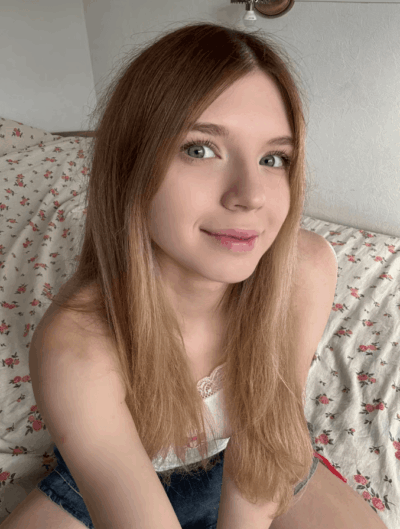 Young woman with long light brown hair and blue eyes sits on a floral bedspread, smiling at the camera.