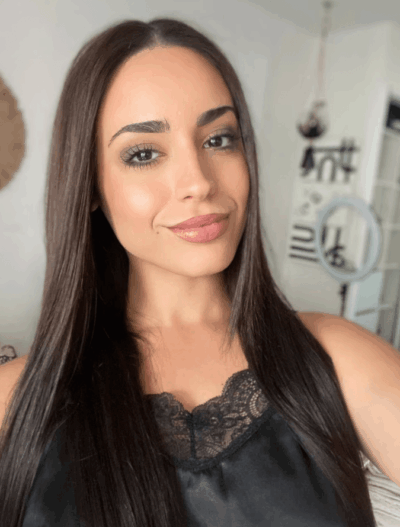 Close-up portrait of a smiling woman with long straight dark hair wearing a black lace-trim top indoors.