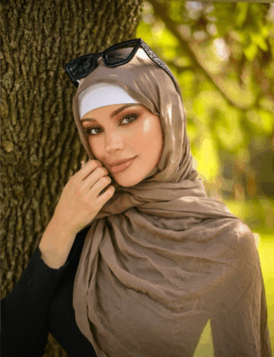 Portrait of a woman wearing a beige hijab and white headband, sunglasses on her head, leaning against a tree in a sunny park.