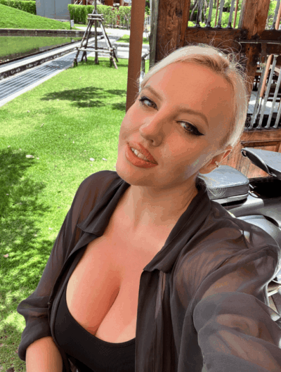 Close-up selfie of a woman with platinum blonde hair wearing a black top and sheer black shirt, outdoors in a sunny garden setting.