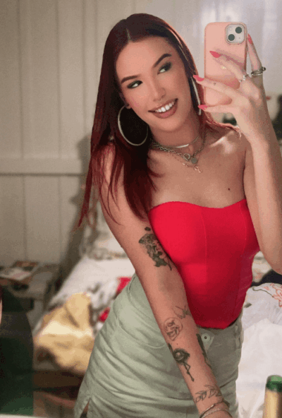 Woman takes a mirror selfie in a bedroom, wearing a red strapless top, light green skirt, with tattoos and hoop earrings and nails painted pink in the photo accessories mold.