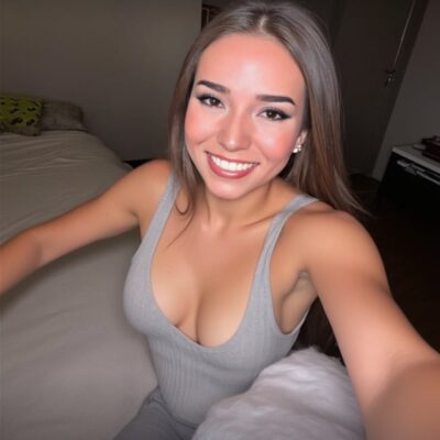 Young woman takes a selfie in a bedroom, wearing a gray sleeveless top and smiling at the camera.