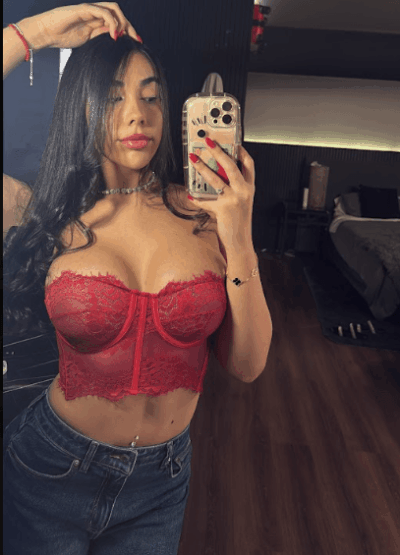 Woman in a red lace bustier taking a mirror selfie in a bedroom, holding her phone with red fingernails visible.