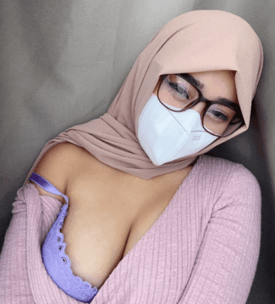 Woman wearing a beige hijab, white mask, and glasses, leaning slightly with a pink knit top and lavender lace bra visible at the neckline in a close-up shot.