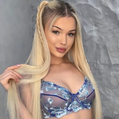Close-up of a smiling woman with long blonde hair wearing a blue floral lace bralette against a gray textured wall.