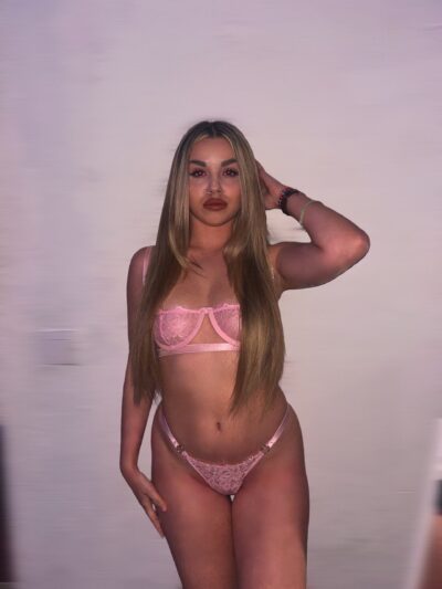 Woman in pink lingerie posing against a light pink backdrop, long straight hair, facing camera with one hand behind head.
