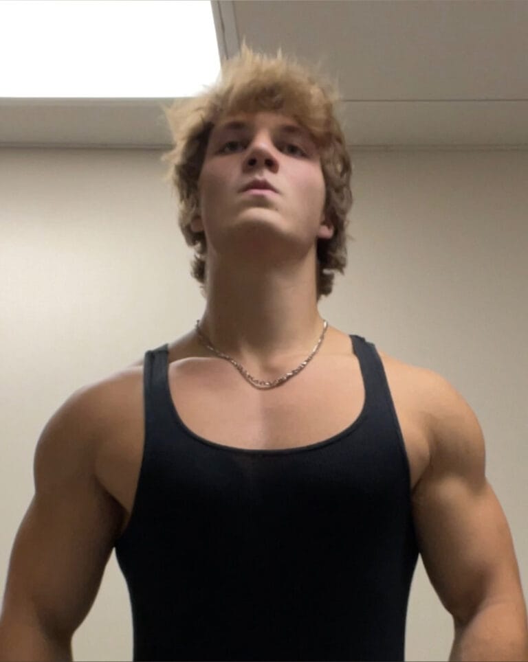 Muscular person in a black tank top stands against a plain wall, viewed from a low angle.