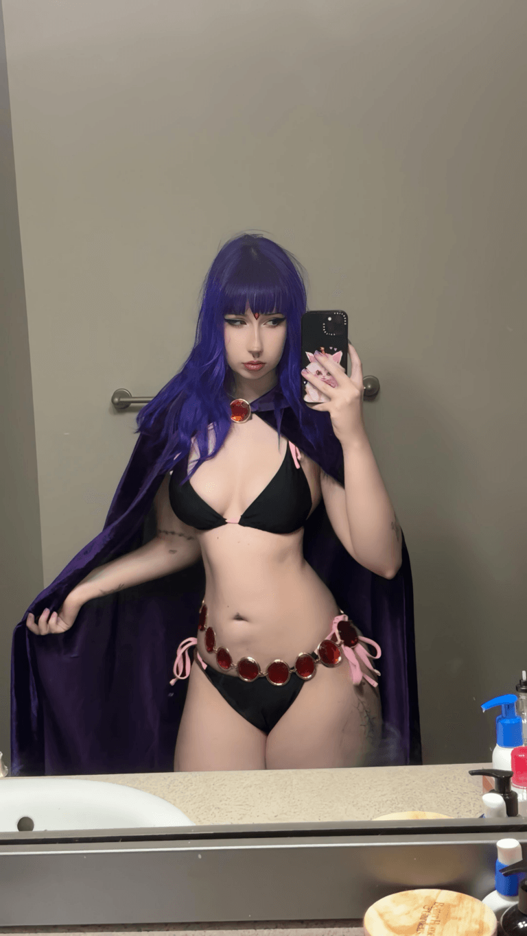 Self-portrait of a person in a purple wig, black bikini, and red gem accessories, wearing a purple cape, taking a mirror selfie in a bathroom.