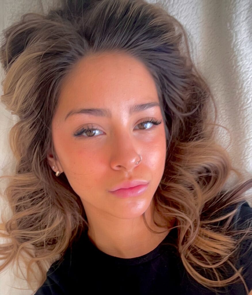 Close-up portrait of a young woman with wavy brown hair, nose ring, hoop earrings, wearing a black top, looking at the camera.