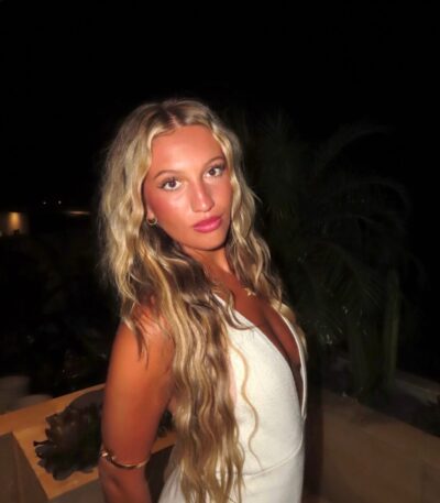 Woman with long wavy blonde hair in a white sleeveless dress poses outdoors at night, softly lit face.