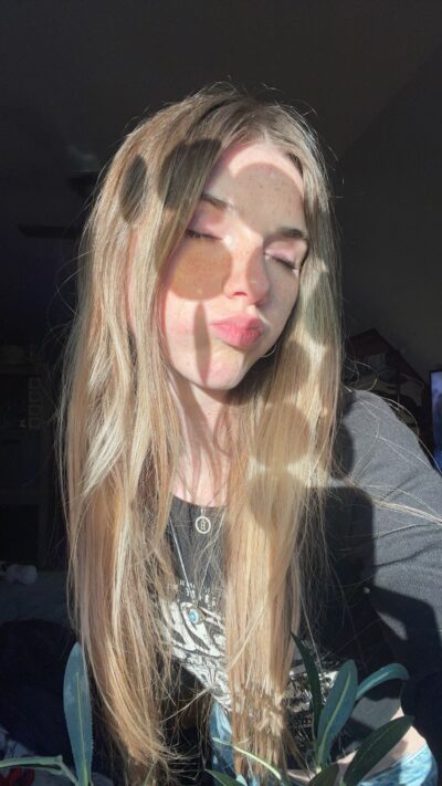Young woman with long blonde hair and freckles sits in sunlight with eyes closed; foreground shows green olive branches partially obscuring her face.