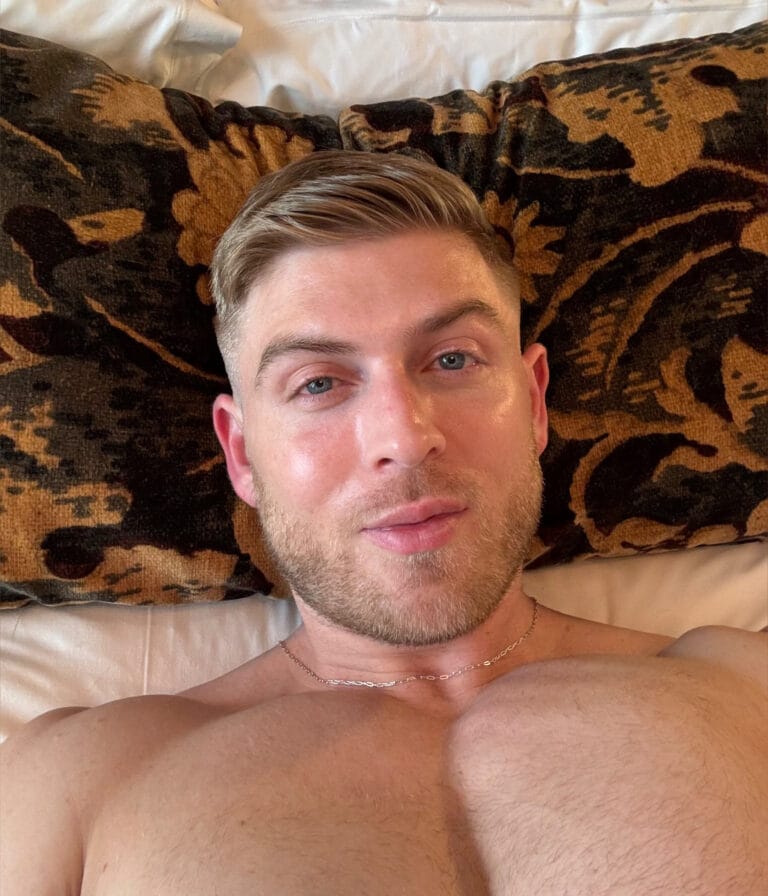 Close-up of a shirtless man lying on a tiger-patterned brown and black blanket, with short, styled hair and a thin chain necklace visible.