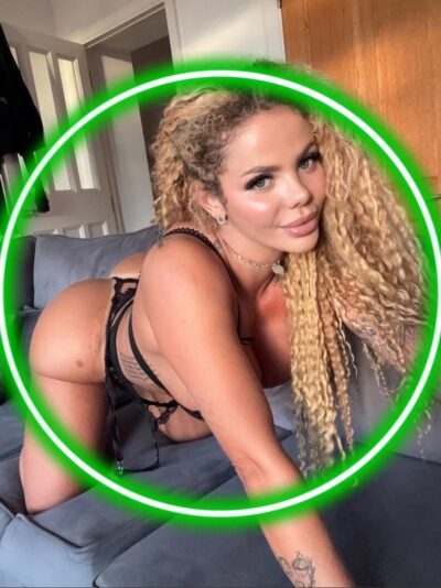 Woman in black lingerie posing on a gray couch, leaning forward with long blonde curls and a green circular glow around her.