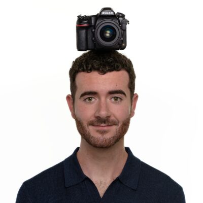 A Nikon DSLR sits on top of a man's head, balanced like a hat against a white background.