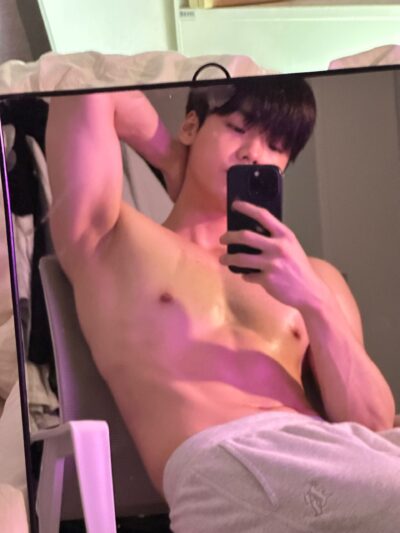 Shirtless man taking a mirror selfie with a smartphone in a bathroom, showing his upper body and arm raised behind the head.