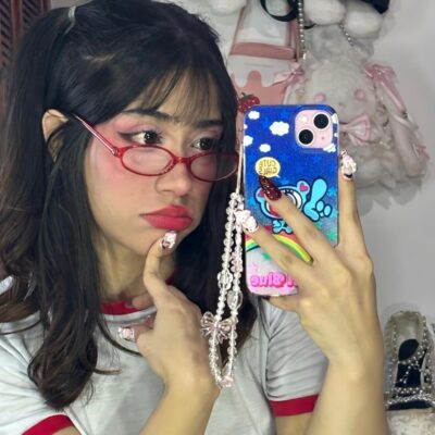 Young woman in red glasses taking a mirror selfie, phone case covered in blue cartoon stickers.