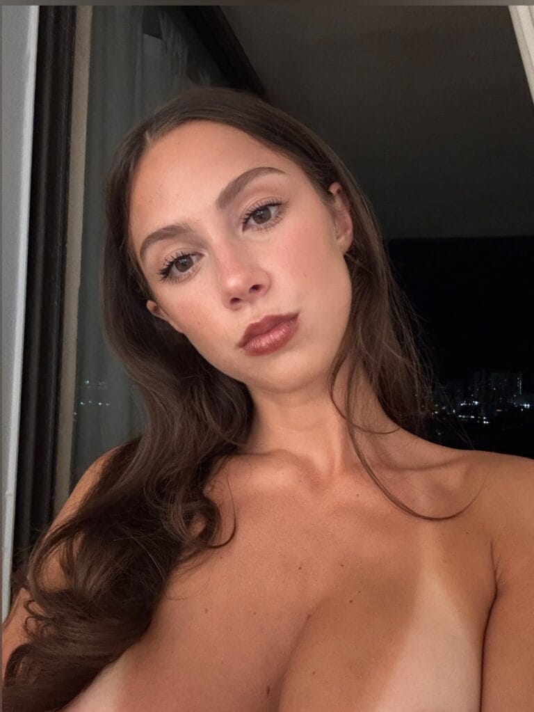 Close-up of a young woman with long brown hair and subtle makeup, bare shoulders, looking at the camera.