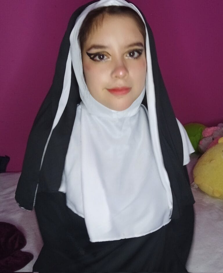 Woman in a black and white nun costume posing for a photo against a pink wall, with makeup and a slight smile.
